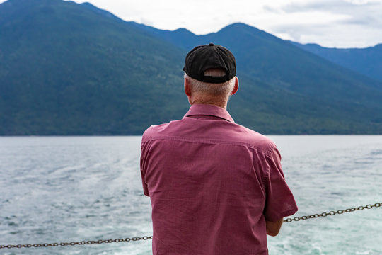 Close Up From Behind Of A Man On The Deck Of A Passenger Ferryboat. Looking Slightly Towards Left At The Lush Mountains Surrounding The Lake.