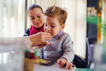 Small little boy three years old child son is sitting in the lap of his mother or aunt while somebody is feeding him having lunch at the table at cafe or restaurant