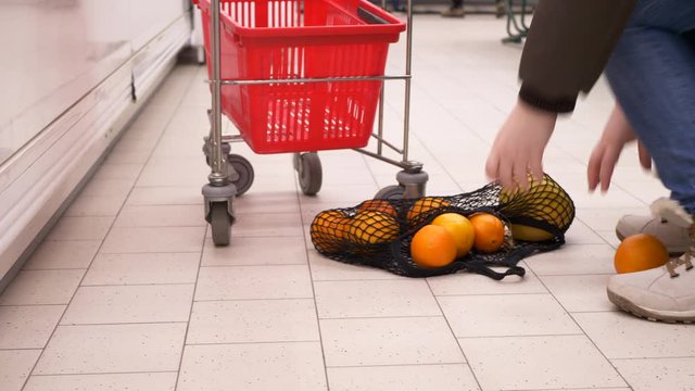 New environmentally friendly packaging for supermarket products. Mesh knitted from natural cotton threads. Adult girl put oranges in net and waving it next to grocery cart