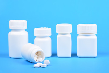 A line of white plastic medicine bottles with one in the foreground and pills spilling out.