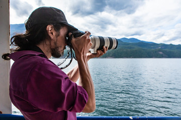 Long haired, young male photographer with an expensive camera with telephoto lens shooting the landscape from the railing of a ferry.
