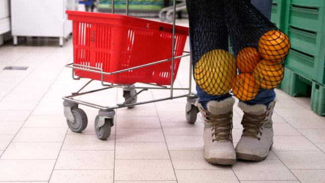 New environmentally friendly packaging for supermarket products. Mesh knitted from natural cotton threads. Adult girl put oranges in net and waving it next to grocery cart