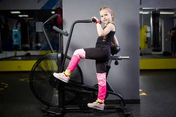 Fototapeta na wymiar Small attractive caucasian child using exercise bike in the gym. Fitness. A little athlete using an air bike for a cardio workout at the crossfit gym. Sport girl sitting on bicycle machine
