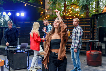 Beautiful girl dancing at foreground while her friends dancing in light of colored lamps during celebration party outdoors.