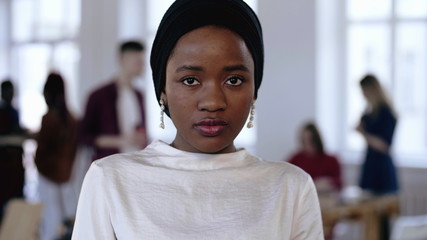 Young serious African business manager woman in ethnic turban looking at camera, smiling at modern loft trendy office.