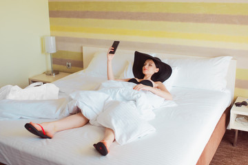 Young girl in a big hat makes selfie on a smartphone while lying in bed. Girl on vacation