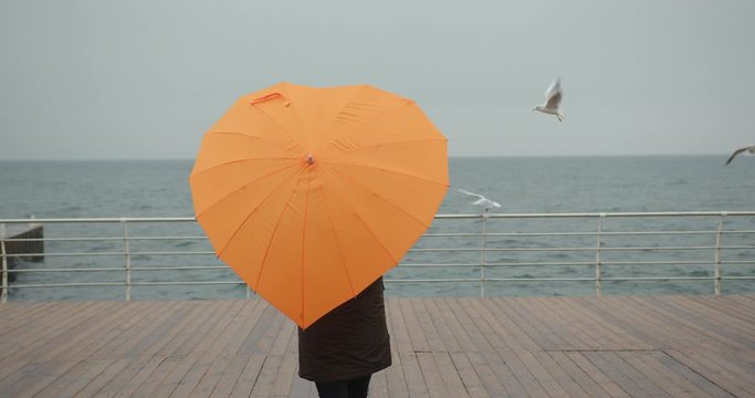 Woman in parka with orange umbrella in heart shape enjoying sea view with seagulls in storm. Overcast weather.