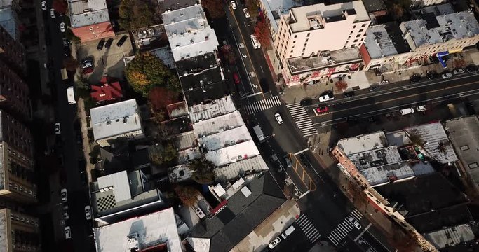 Cars, trucks, and taxis pass on the streets and intersections of New York City past apartment buildings and pedestrians casting shadows. Aerial drone