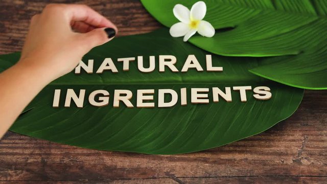 Natural Ingredients message on tropical banana leaf with hand placing monoi frangipani flowers around it