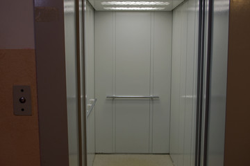 The Elevator and its interior painted light gray with metal handrails and overhead lighting visible through fully open doors against a peach colored wall in an multistory apartment building