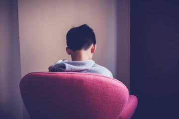Sad preteen tween mixed Asian boy sitting alone in chair facing wall, depressed,teen Autism...