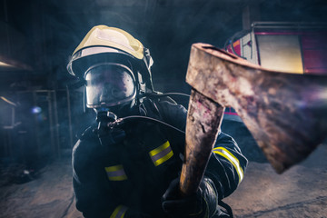 Portrait of a female firefighter while holding an axe and wearing an oxygen mask indoors surrounded...