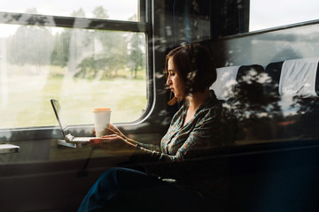 A middle aged woman sitting by the window of a commuter train with a laptop and coffee in a...