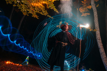 Fototapeta premium Sorcerer (magician) casting a spell while standing in the magical woods. Holding his magical staff. Magic illumination. Autumn holiday celebration. Mystery and nightmare concept.