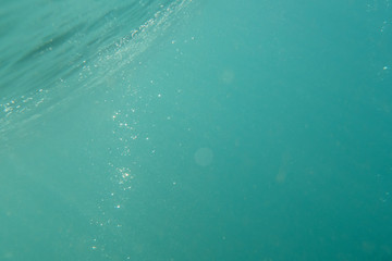 Light rays underwater beneath the surface of a turquoise breaking ocean wave, as bubbles rise to...