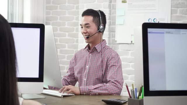 Young Asian telemarking agent wearing microphone headset talking to customer in call center office  