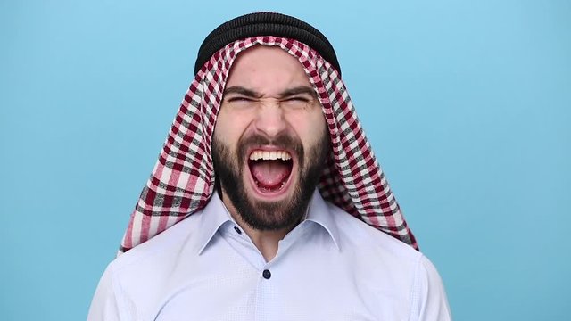 Bearded young arabian muslim man 20s in keffiyeh kafiya ring igal agal casual clothes posing isolated on pastel blue background. People religious lifestyle concept Screaming angry frustrated irritated