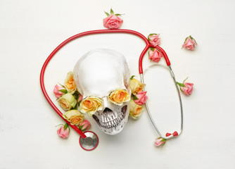 Stethoscope with flowers and human skull on white wooden background