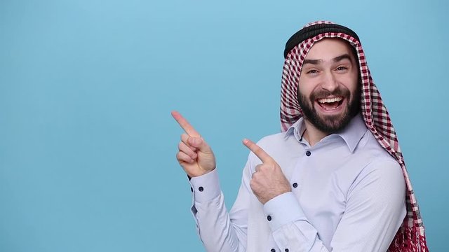 Bearded young arabian muslim man 20s in keffiyeh kafiya ring igal agal casual clothes posing isolated on pastel blue background. People religious lifestyle concept Pointing fingers on copy space place
