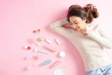 Obraz na płótnie Canvas Smiling Asian woman lying on pink background with her cosmetics tools. Top view