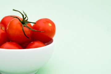 Little fresh red uncooked tomatoes with a green sprig in a white bowl on light green background. Healthy eating and raw food concept.