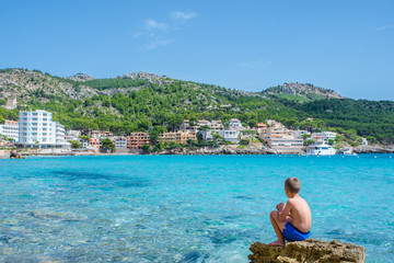 a boy sits on a stone near the blue Mediterranean Sea with his back to us, Mallorca, Spain