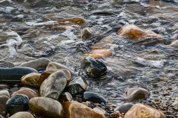 Large and smooth stones in the coastal waves of salty sea water. The coast of the black sea foams and roars, beating against a huge pebble, illuminated by sunlight