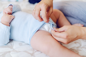 Close up of caring mother sitting on the bed in bedroom, changing baby buckling bodysuit. Bedroom interior.