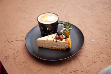 A black cup with a coffee is on a black plate near a piece of cake on which there are nuts and fruits