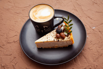 A black cup with a coffee is on a black plate near a piece of cake on which there are nuts and fruits