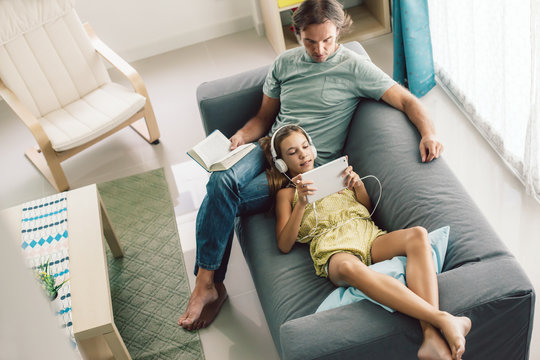 Father and pre teen daughter playing tablet and reading book while relaxing together on couch in room at home