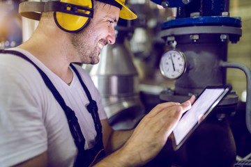 Smiling dedicated factory worker in overalls, with hardhat and antiphons using tablet for checking on boiler. Power plant interior.