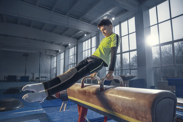 Fototapeta na wymiar Control. Little male gymnast training in gym, flexible and active. Caucasian little boy, athlete in sportswear practicing in exercises for strength, balance. Movement, action, motion, dynamic concept.