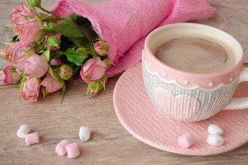 Coffee cup on a wooden table with bouquet of pink roses. Knitted heart shaped cup