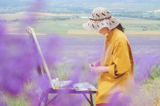 Girl artist is painting lavender field on the plein air. Box easel with canvas, oil paint, palette, and tools of an artist.