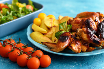 closeup delicious chicken wings with potatoes, vegetable salad, red tomatoes on blue plates and background
