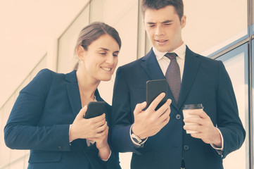 Puzzled manager with takeaway cup showing smartphone screen to female coworker. Two colleagues with phones sharing news during coffee break. Sharing news concept