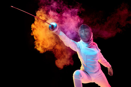 Contrasts. Teen girl in fencing costume with sword in hand isolated on black background, neon lighted smoke. Practicing and training in motion, action. Copyspace. Sport, youth, healthy lifestyle.