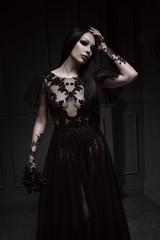Beautiful brunette woman with gothic makeup wearing stylish black dress and posing on grey...