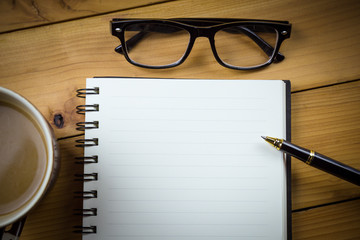 Blank notebook with pen and with glasses next to cup of coffee on wooden table,business concept.