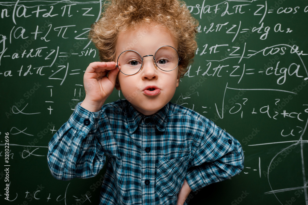 Wall mural smart kid touching glasses and standing with hand on hip near chalkboard