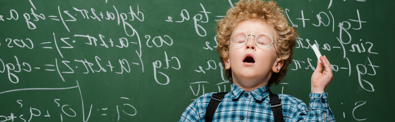 panoramic shot of curly kid in glasses sneezing near chalkboard with mathematical formulas