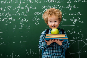 cheerful kid holding books and apple near chalkboard with mathematical formulas