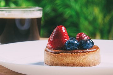 Closeup of beautiful mini tart with berries and cup of coffee. Delicious pastry with strawberries and blueberries on plate on table with blurred green view in background. Breakfast concept.