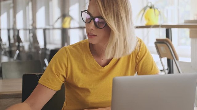 An attractive young woman wearing eyeglasses is making notes on her notebook while working on her laptop in a conference hall