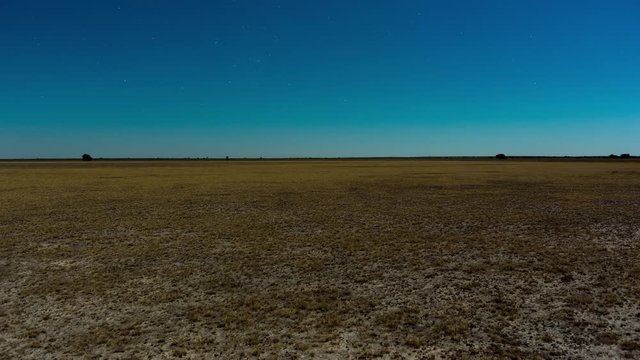 Static vast open arid moon light African landscape timelapse while Milky Way sets against blue sky in Southern Hemisphere, with scattered grass patches in drought season, Botswana, Central Kalahari.