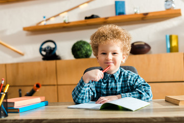 Naklejka premium happy and curly kid holding pen near notebook and books on desk