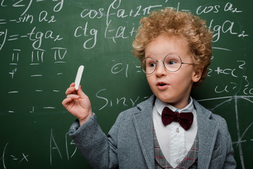 smart child in suit and bow tie holding chalk near chalkboard with mathematical formulas