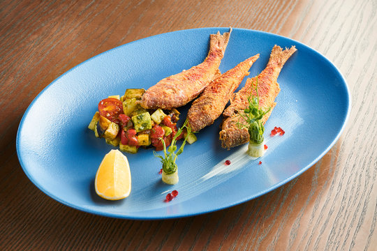 Deep-fried red mullet fish on a blue ceramic plate on a wooden background. Restaurant serving fish with salad. Close up view on tasty seafood. Film effect during post. Soft focus