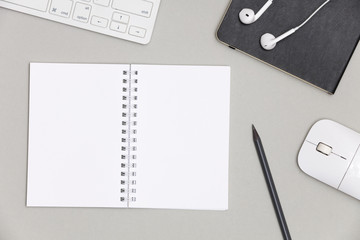 A blank white notebook for notes with a pencil on a gray background surrounded by a keyboard, headphones and mouse. Creative flat lay photo of workspace desk with copy space. 